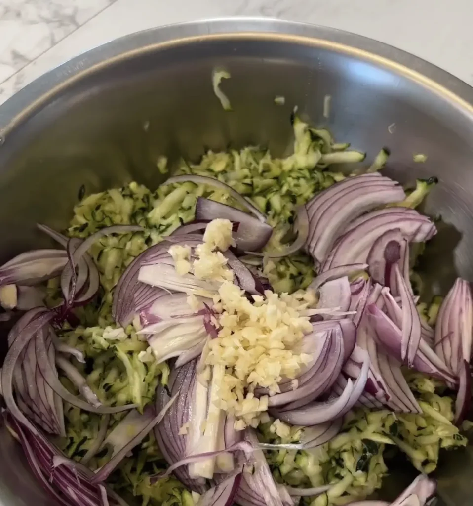 A large bowl, combining the drained zucchini, sliced onion, minced garlic, and chickpea flour.